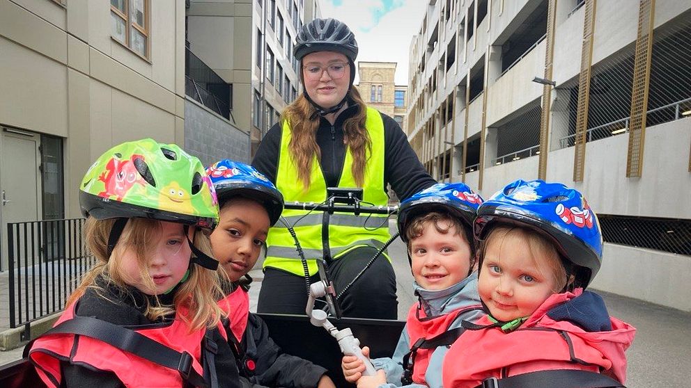 Flera barn med cykelhjälmar färdas i en lådcykel, vuxen med reflexväst cyklar bakom.