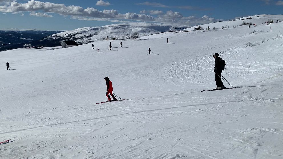 Skidåkare på en snöklädd backe i fjällen under en klarblå himmel, med flera personer utspridda i bakgrunden som åker skidor.