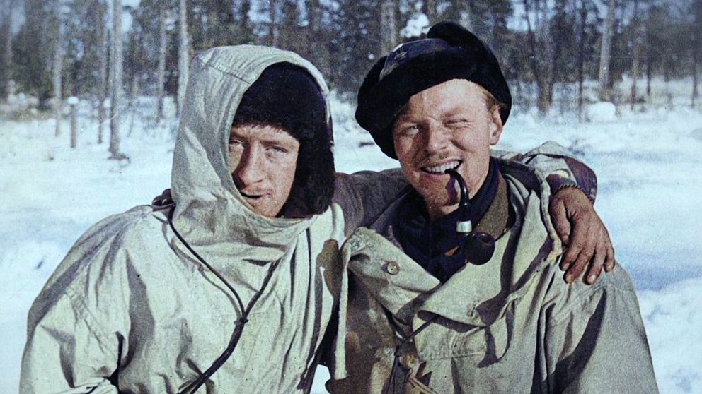 Två personer i vinterkläder står tillsammans i ett snöigt skogslandskap.