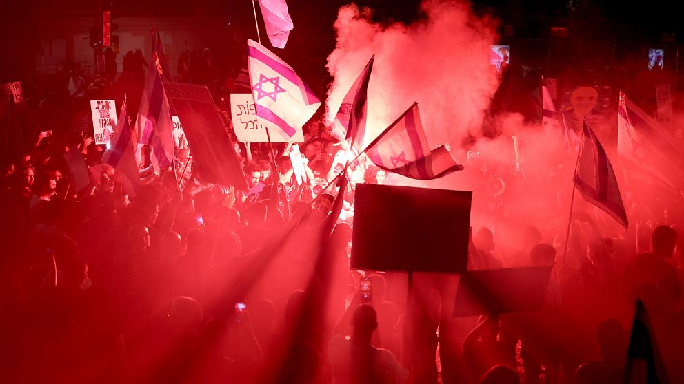 Demonstranter i Tel Aviv upplyst i rött ljus och rök viftar med israeliska flaggor och skyltar.