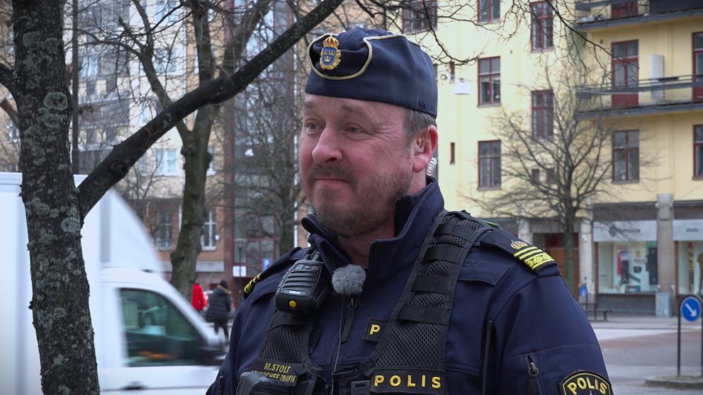 En polis står i en stad, med byggnader och ett vitt fordon i bakgrunden.