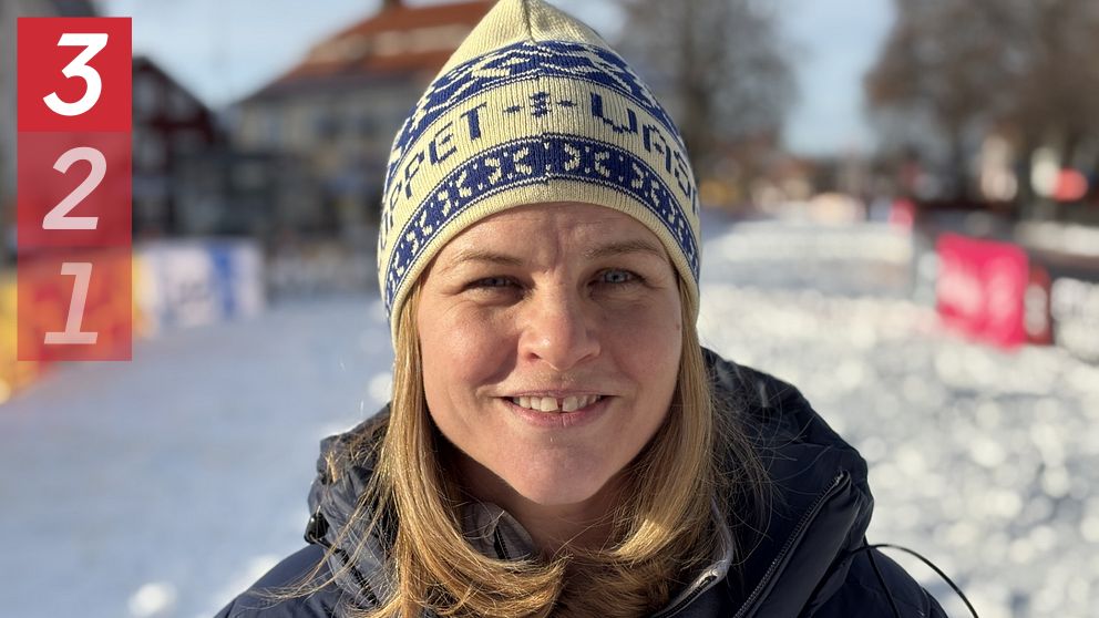 Person i vinterjacka och mössa står på en snöklädd gata med hus i bakgrunden.