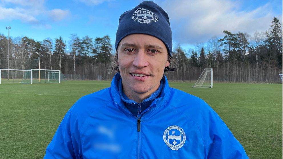En person i en blå sportsjacka och mössa står på en fotbollsplan, omgiven av träd och två fotbollsmål i bakgrunden. Det är en klar dag.