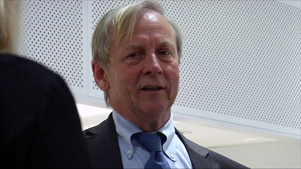 En man i kostym sitter framför en person som står vänd mot honom. De befinner sig i en modern inomhusmiljö med vita väggar och en minimalistisk inredning. Samtalet verkar fokuserat och allvarligt.