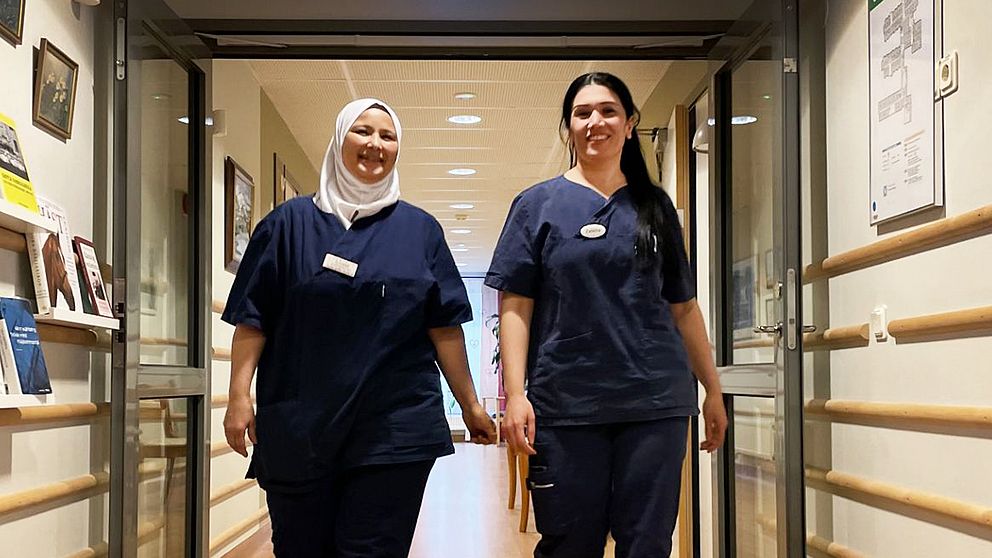 Kvinnorna Ilham och Zalikha i blå arbetskläder går i en korridor på ett äldreboende i Säffle