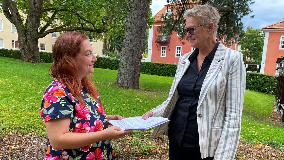 Anna Ekman-Brescia, en kvinna med blommig blus och rött hår, lämnar över en namnlista till förvaltningschefen för tandvård i Dalarna, en äldre kvinna med randig kavaj.