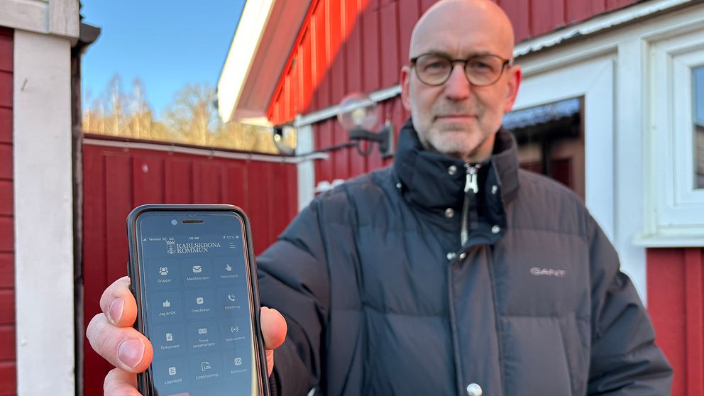 Person, Anders Viklander, i svart jacka visar mobiltelefon som har en krisinformationsapp installerad mot kameran utanför rött hus.