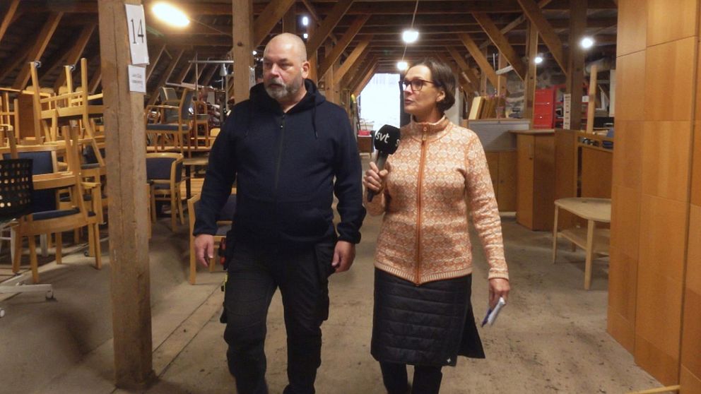 En man och en kvinna går tillsammans genom ett stort förråd, omgivna av stolar och bord. Kvinnan håller en mikrofon och talar. En stor, öppen lokal med träbjälkar i taket.