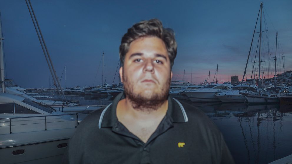 Olof K Gustafsson i mörk tröja står vid småbåtshamn i skymning. Båtar och mastar syns i bakgrunden.