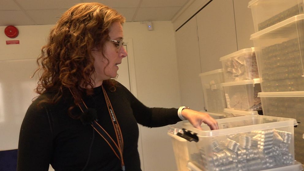 En person står vid ett bord i ett rum fyllt med förvaringslådor, och räcker ner en hand mot en öppen plastbehållare med tablettkartor.