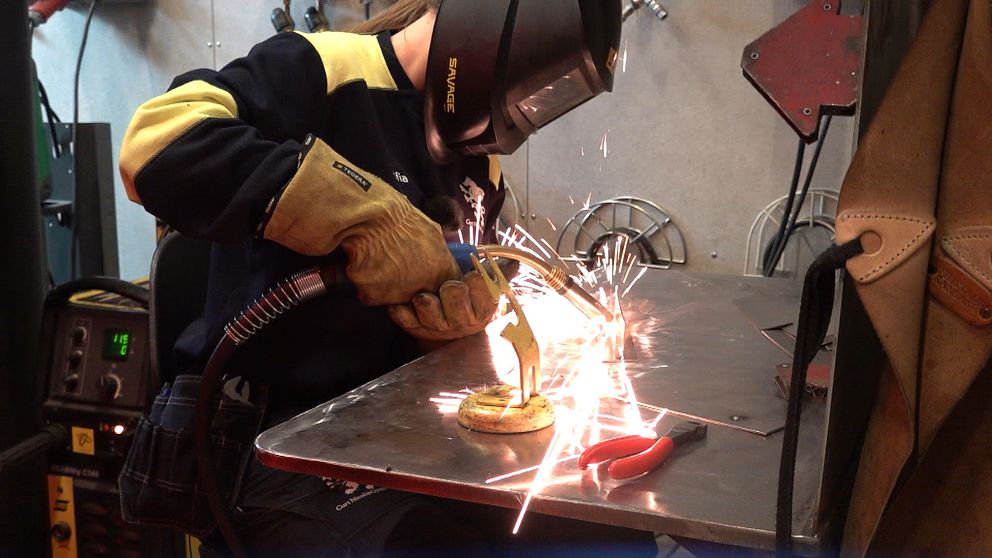 En person svetsar på ett metallstycke, vilket skapar gnistor. Personen bär skyddshandskar och en svetsmask, och arbetar på en metallplatta som är placerad på ett arbetsbord. I bakgrunden syns verktyg samt svetsutrustning.