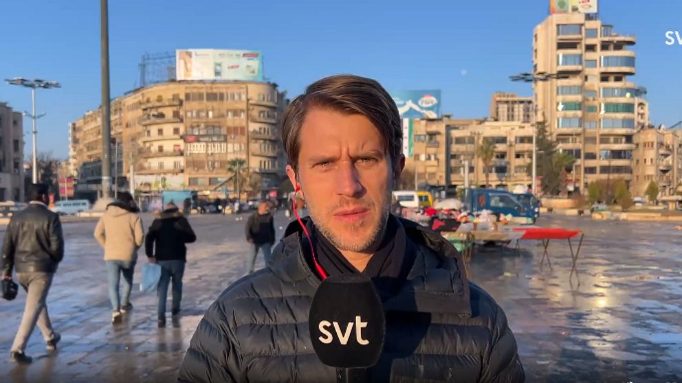 Thomas Thoren står på ett torg och rapporterar med byggnader i bakgrunden