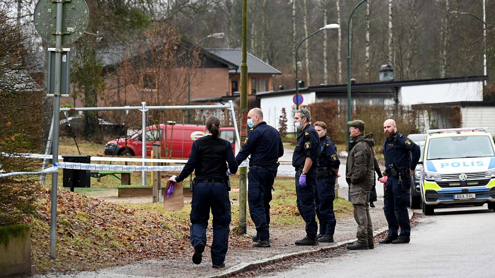 Flera poliser och en civilperson står vid en brottsplats som är avspärrad med polisband. En polisbil är parkerad på vägen.