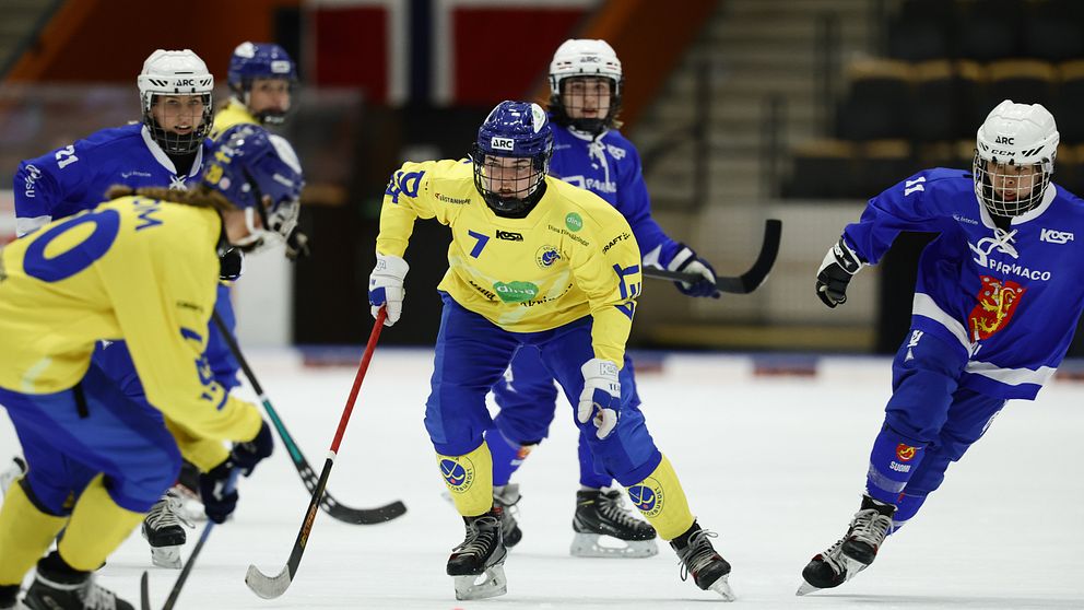 Svenska och norska bandyspelare tävlar på isen med Norgeflaggan i bakgrunden.