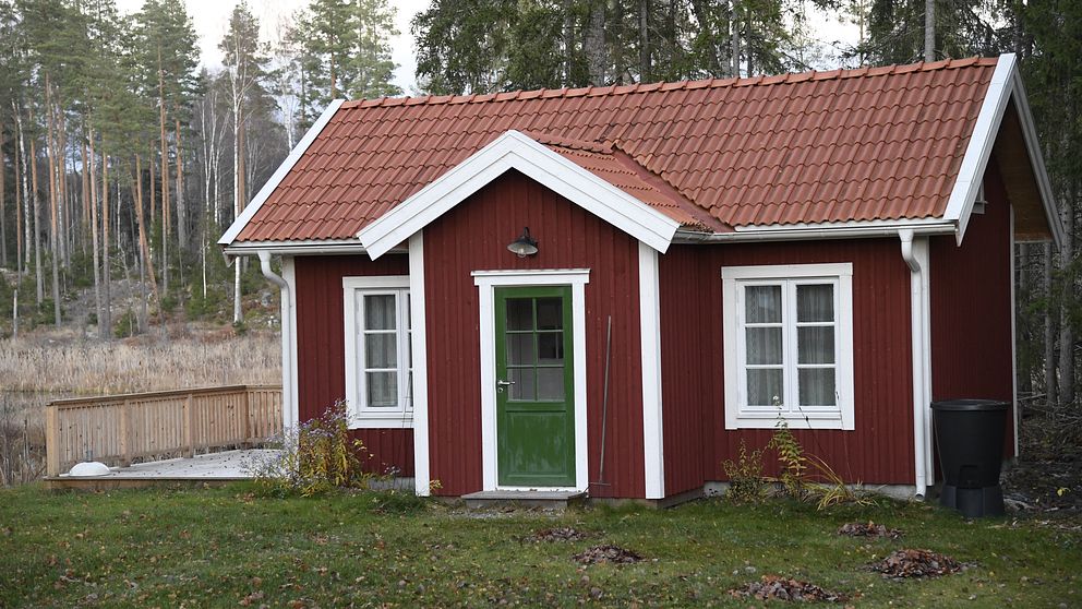 Röd stuga med vita knutar och gröna dörrar, omgiven av skog och gräsmatta.