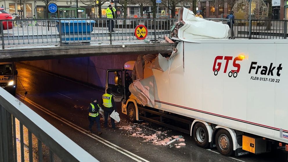 Lastbil från GTS Frakt har fastnat under en bro och fått sin trailer skadad, med delar av taket sönder. Två personer i reflexvästar inspekterar skadan och försöker lösa situationen. En skylt på bron visar höjdbegränsningen på 3,4 meter.