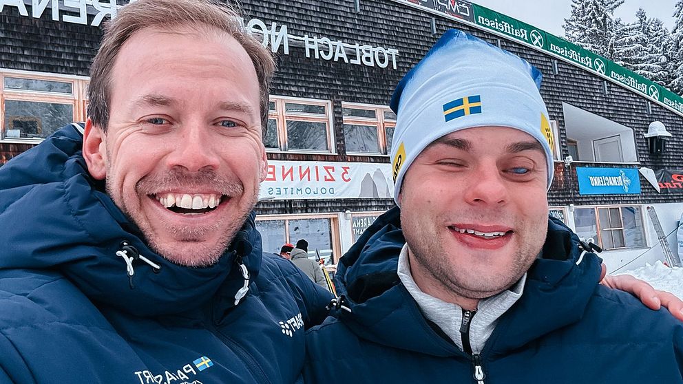 Två personer i vinterkläder står utomhus framför en byggnad med skyltar på ett främmande språk. Det är snö på marken och taket, vilket tyder på kallt väder.
