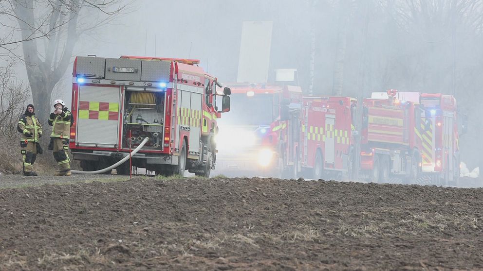 Brandbilar och brandmän vid rökfyllt område på landsväg nära åker.
