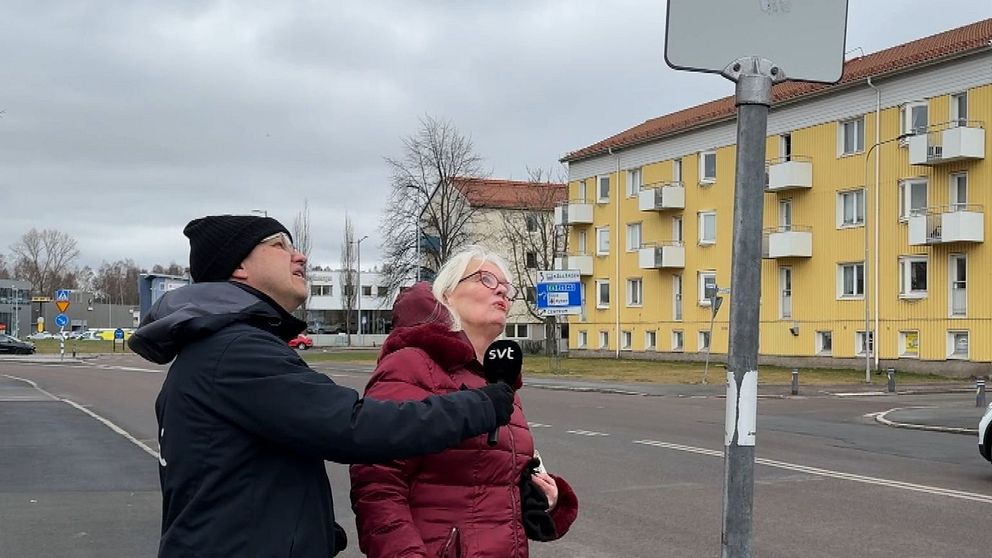 En man och kvinna tittar på en busskylt vid en gata med ett gult bostadshus i bakgrunden.