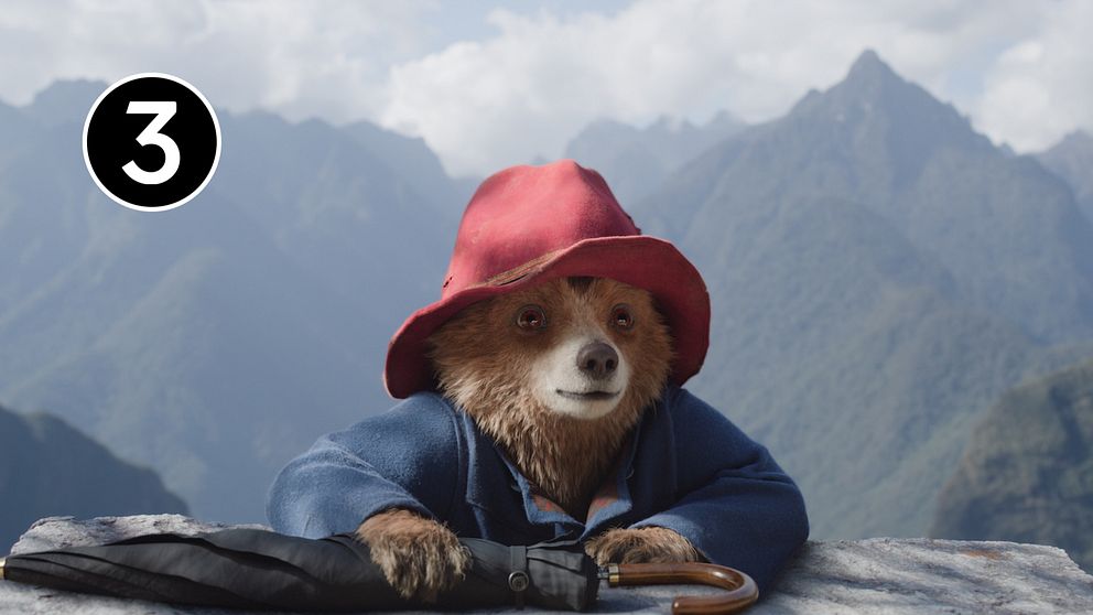 Paddington i en stor röd hatt och blå kappa sitter vid en bergskant med en storslagen vy av fjäll i bakgrunden.