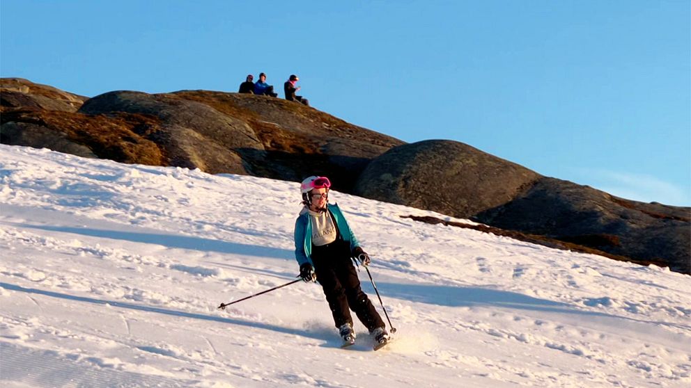 Skidåkare i solnedgång, backen omgiven av berg och blå himmel. Tre personer på toppen.