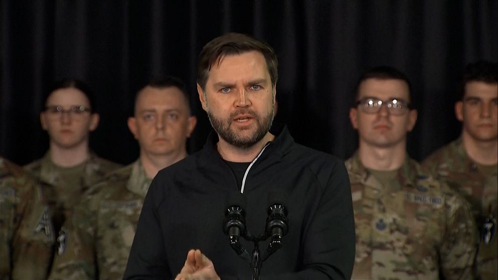 Talare står framför militär personal. Mörk bakgrund.