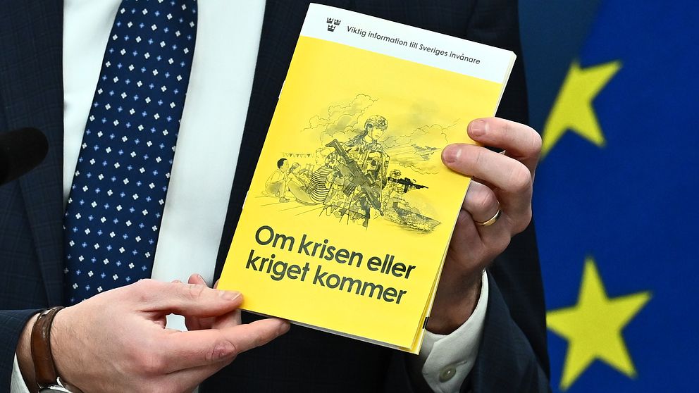 En person håller upp en bok med titeln ”Om krisen eller kriget kommer”, som har en gul omslag med en illustration i gråtoner. I bakgrunden syns en EU-flagga. Boken verkar vara relaterad till krisberedskap och säkerhet.
