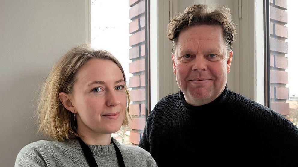 En kvinna med kort, ljust hår och en grå tröja står bredvid en man med mörkt hår som bär en svart tröja. De står vid ett fönster med ljusinsläpp och en bakgrund av tegelvägg.