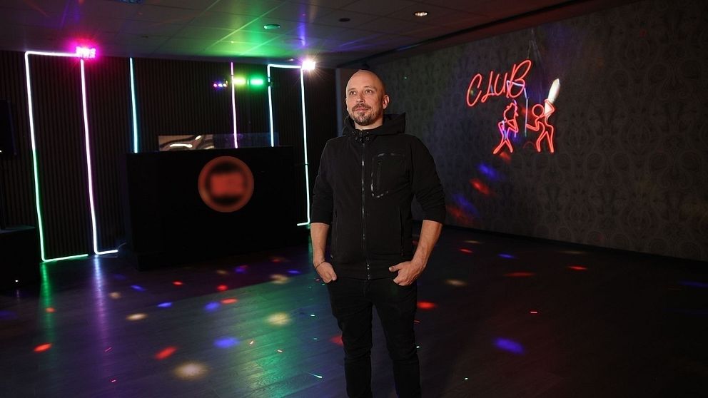 En person står i ett mörkt rum med färgglad belysning, inklusive blinkande ljus och neonskyltar. Rummet har en DJ-bås med en rund logotyp synlig. På väggen finns en neonskylt med illustrationer av dansande figurer. Atmosfären ger en känsla av nattliv och fest.
