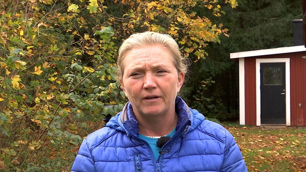 Personen bär en blå jacka och står framför en skogsmiljö med höstlöv i bakgrunden. En röd stuga är synlig i bakgrunden.