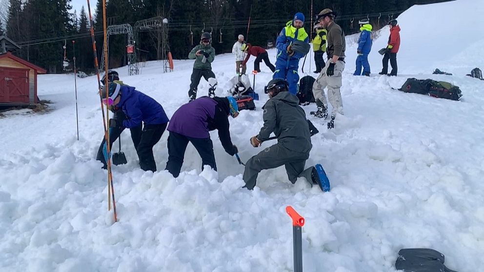 En grupp människor iförda vinterkläder deltar i en gemensam snöaktivitet i en skidbacke, med några som gräver i snön och andra som observerar. De omges av vinterlandskap med skidliftar i bakgrunden.