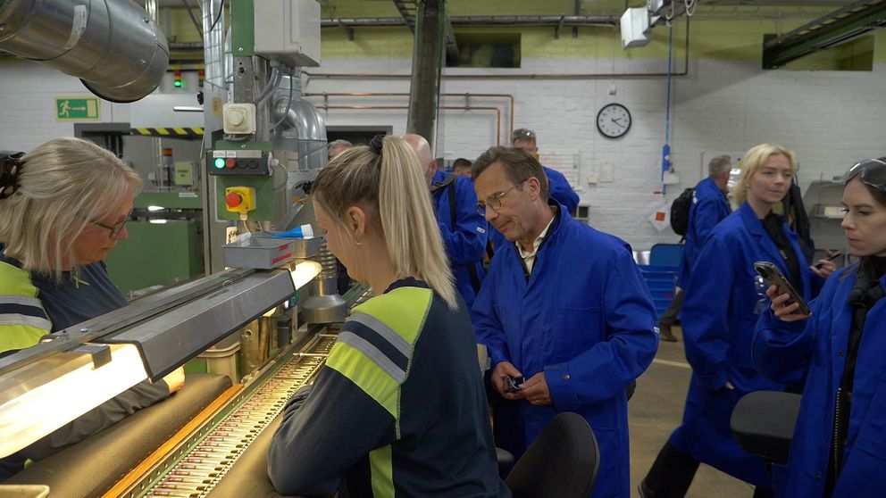 Personer i blå arbetskläder besöker en industrihall och studerar en pågående produktion.