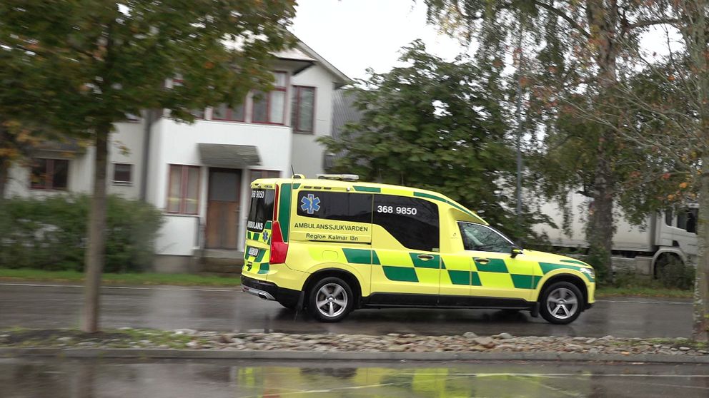 Ambulans på regnvåt asfalt. Träd och ett vitt hus i bakgrunden.