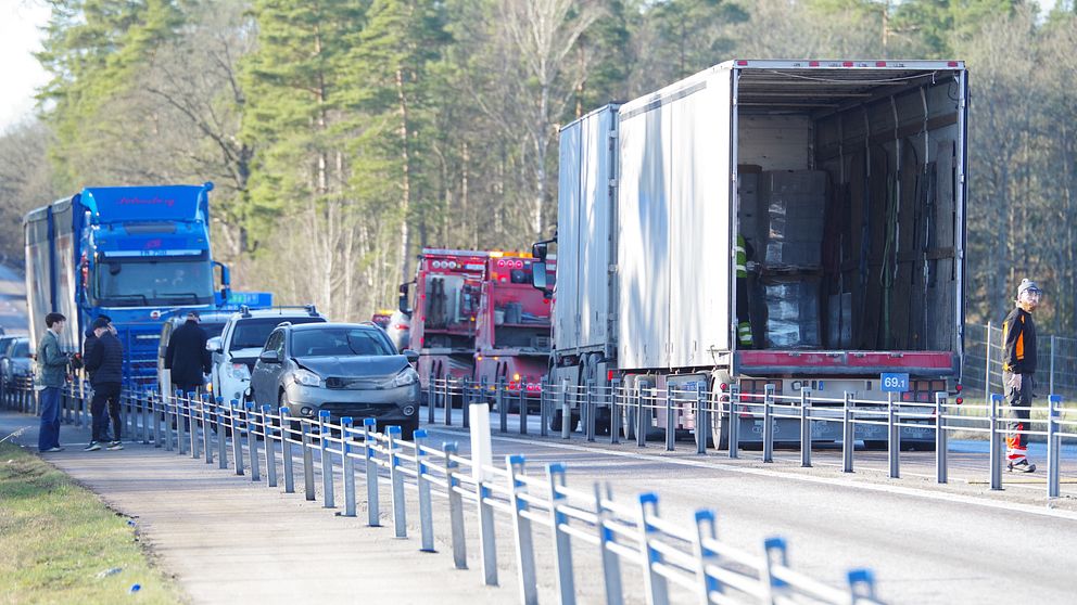Trafiksituation på en landsväg med flera lastbilar och en personbil stannade på vägrenen. En lastbil har bakdörren öppen, och människor står i närheten, förmodligen involverade i en olycka eller transportrelaterad situation.
