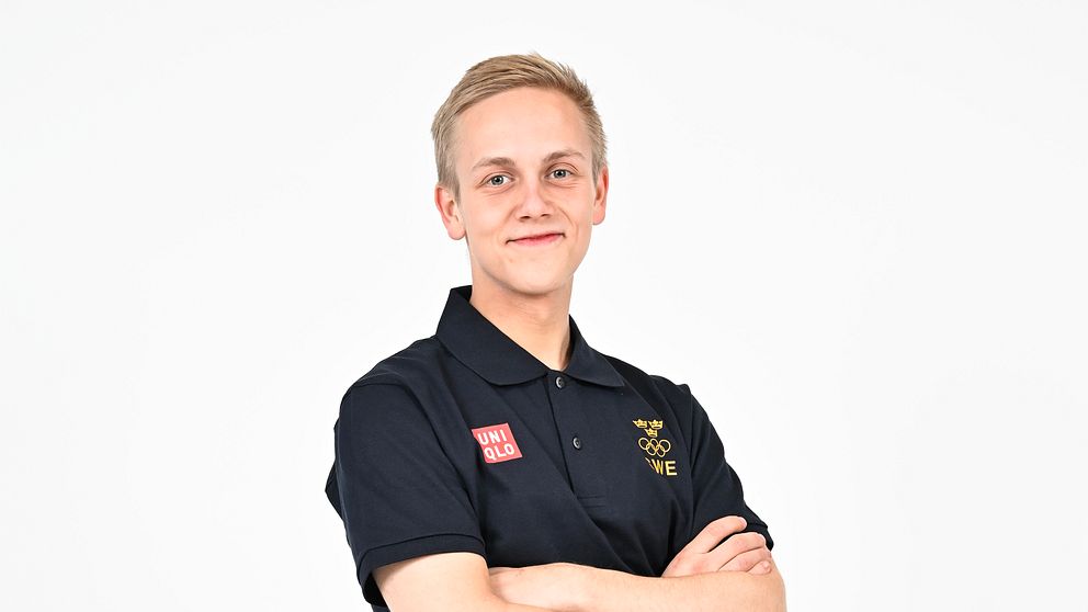 Blond ung man iklädd blå pikéskjorta med OS-ringarna tittar in i kameran och ler.