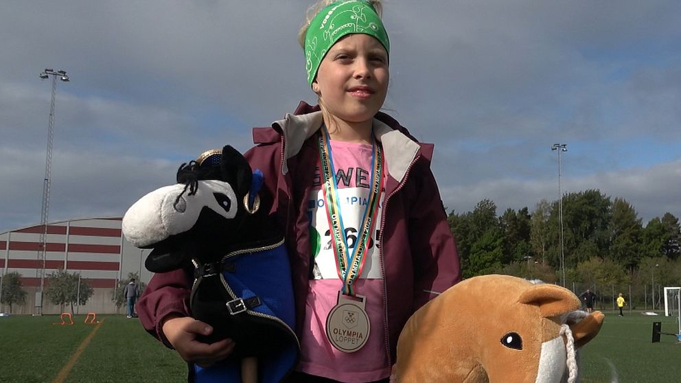 Bilden visar flicka med medalj runt halsen och två käpphästar