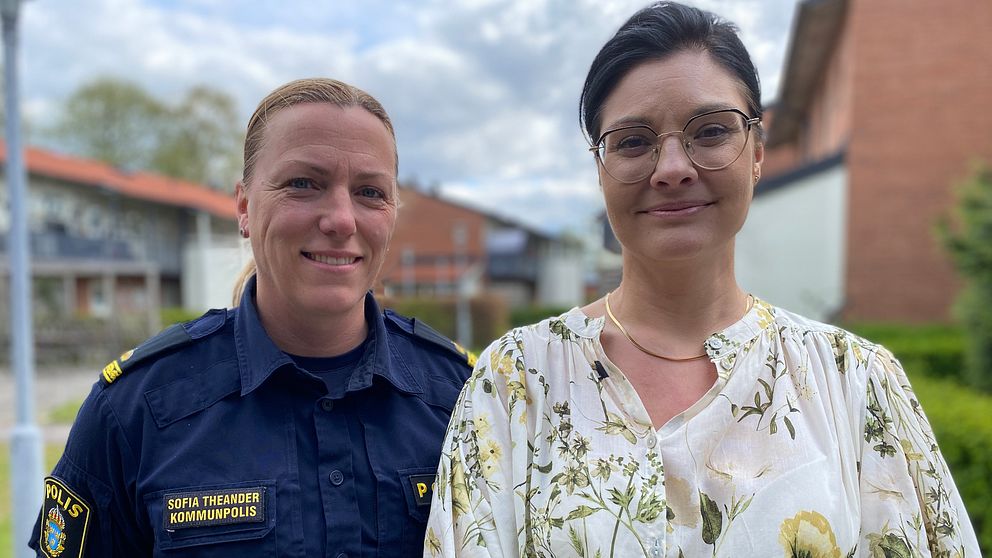 Två personer står utomhus, en i polisuniform och en i mönstrad blus. Hus syns i bakgrunden.