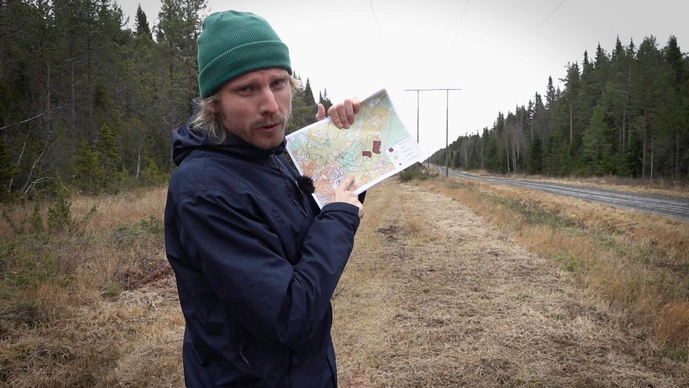 SVTs reporter står längs ett motionsspår omgiven av skog, hållande en karta och pekar på den. I bakgrunden syns kraftledningar som sträcker sig längs vägen.