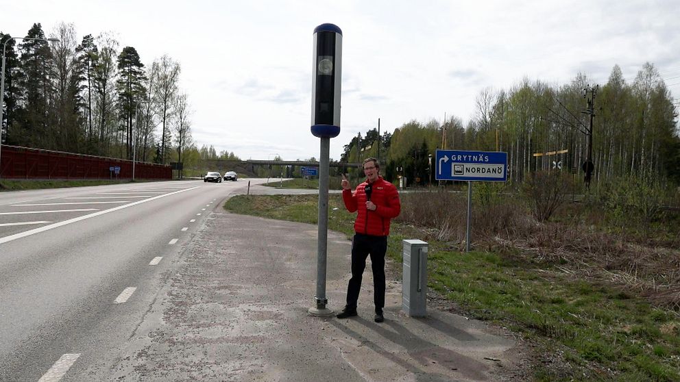 Reporter står med röd jacka och pekar på en fartkamera intill en väg.