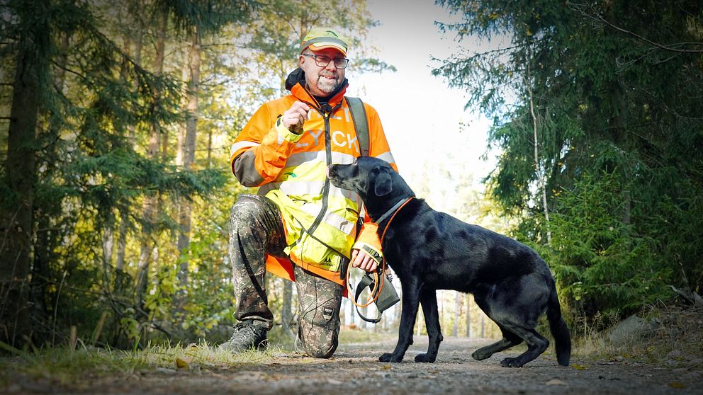 En person i orange och gul arbetsjacka står på knä i en skogsmiljö och interagerar med en svart hund som står bredvid. Området är omgivet av träd och ger intryck av att vara en friluftsmiljö, där personen verkar ta en paus för att umgås med hunden.