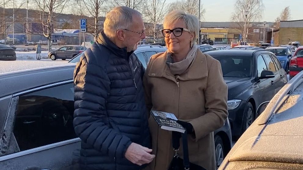 Två personer står nära varandra på en parkeringsplats, omgivna av bilar. En av dem håller en bok, och det är vinter med snö på marken. I bakgrunden syns träd och en byggnad.