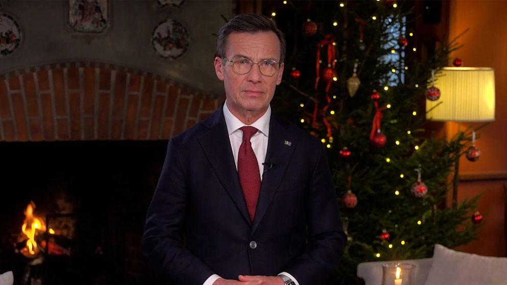 Ulf Kristersson står framför en tänd brasa, medan en julgran lyses upp i bakgrunden. Stämningen är varm och julig.