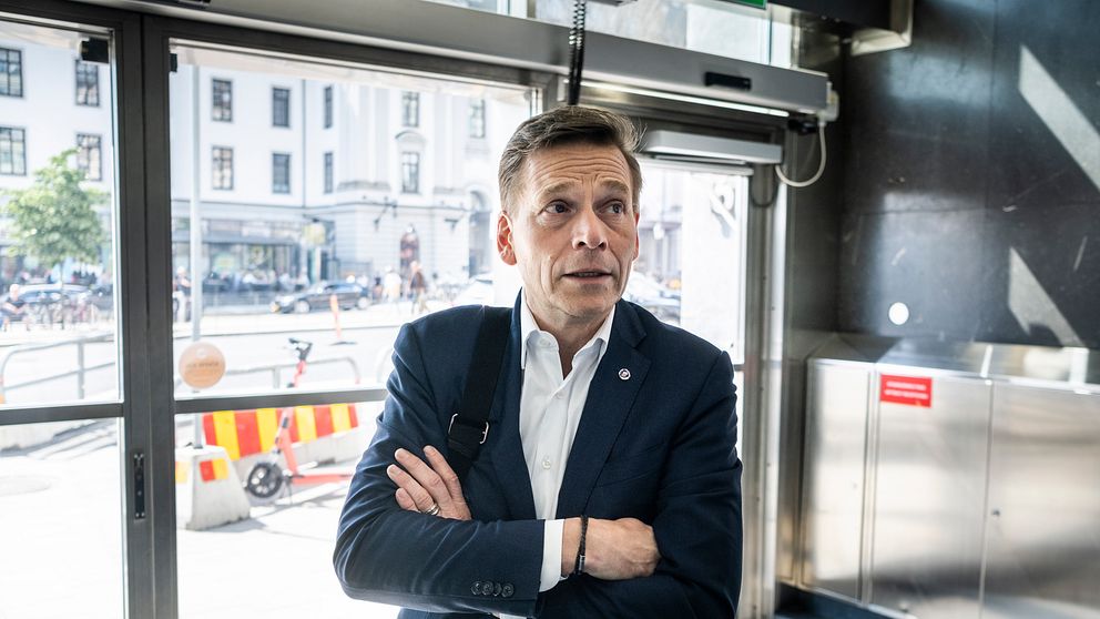 En man i kostym står med korsade armar inomhus vid en glasdörr mot stadsmiljö.