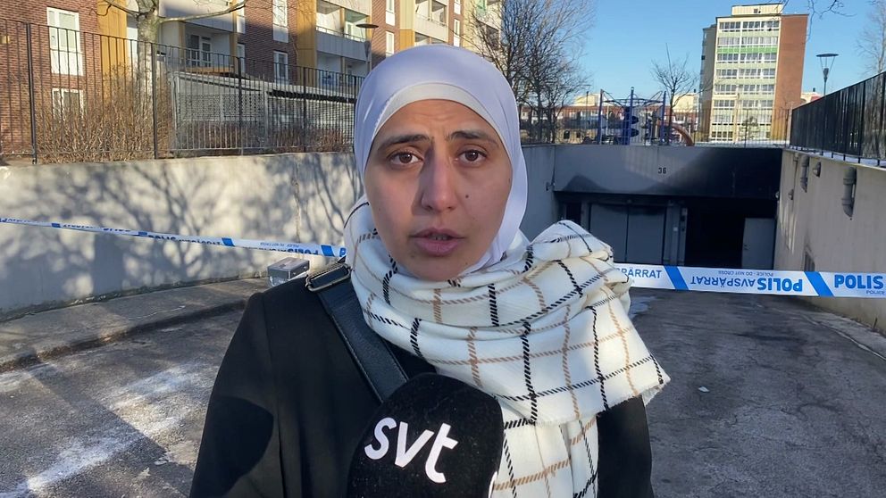 En person iförd en svart jacka och vit- och svartmönstrad sjal står framför ett avspärrat område med polistejp. Bakgrunden visar en porten till ett underjordisk garage, bostadshus och en klarblå himmel. En mikrofon med SVT:s logotyp hålls av personen.