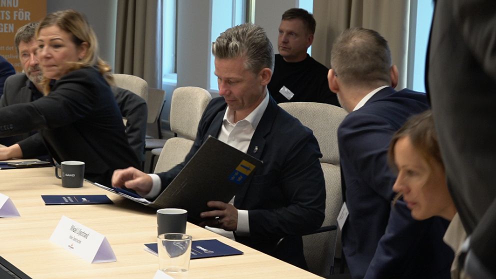 En grupp personer sitter runt ett konferensbord och diskuterar, med mappar och kaffe framför sig. Namnskyltar är placerade på bordet.