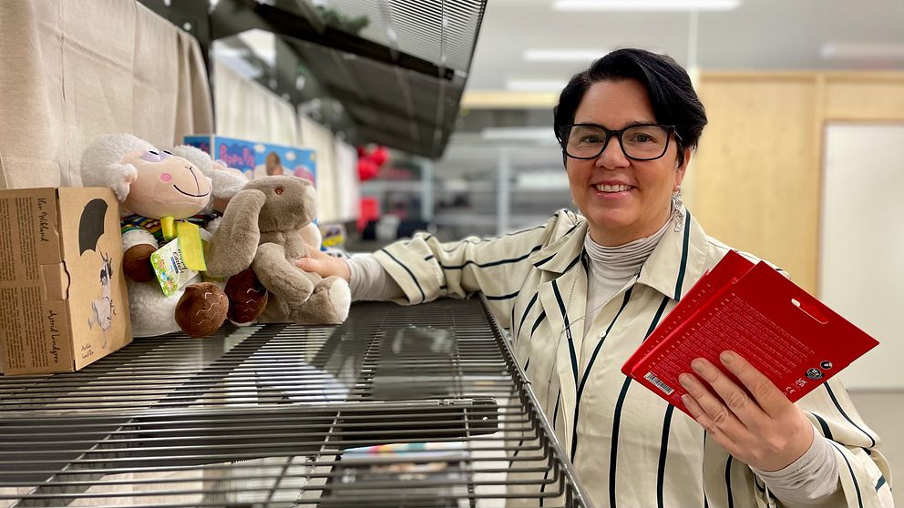 En person står vid en lagerhylla i en butik, holding flera röda kort. På hyllan finns mjukisdjur och förpackningar. Bakgrunden är otydlig med hyllor och produkter synliga.