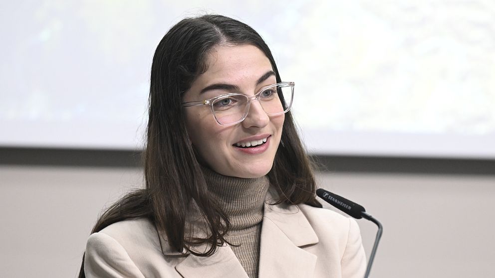 Kvinna i ljus kavaj står vid en mikrofon framför en presentationsduk.