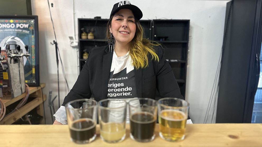 En kvinna med en t-shirt med texten ”Sveriges oberoende bryggerier” och svart kavaj och keps står bakom en bardisk.