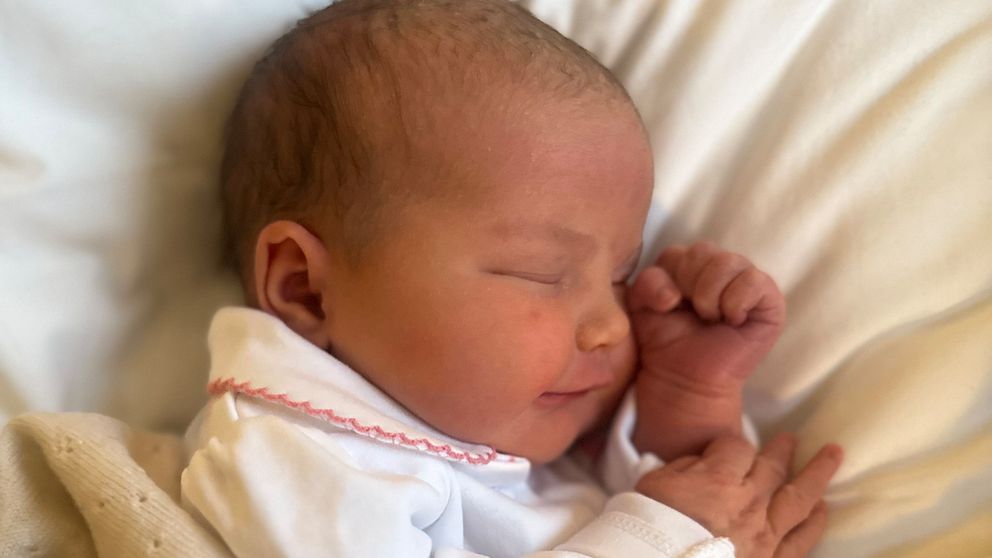 En nyfödd baby insvept i en beige filt, klädd i en vit pyjamas med rosa detaljer, ligger på en vit kudde.