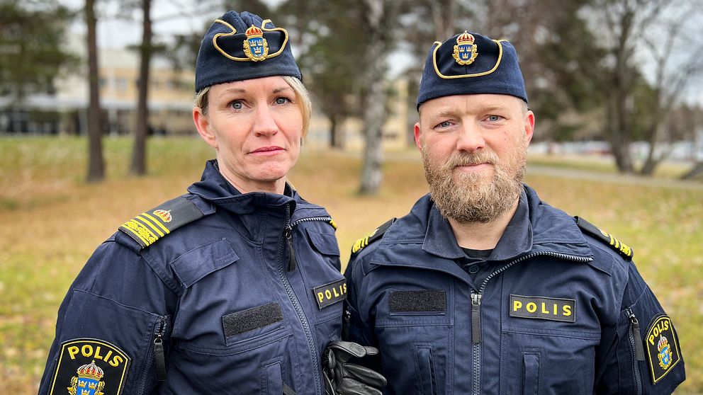 Två poliser poserar tillsammans utomhus, klädda i mörkblå uniformer med svenska polisens insignier. Bakgrunden visar en park med träd och en byggnad i fjärran, vilket antyder en lugn miljö.